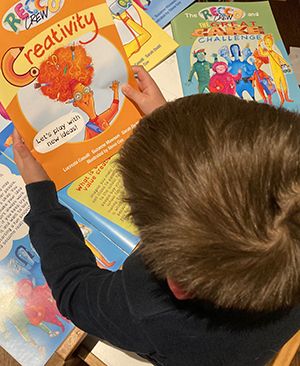A young child with short brown hair is seated at a table, looking down at and holding a colorful educational booklet titled 'Creativity,' surrounded by several other bright, cartoon-style activity booklets spread out across the surface.